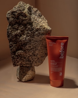 A large piece of rough-textured rock stands beside a red-orange tube of toothpaste labeled 'before' and 'purifying toothpaste'. The setting is neutral-toned with soft lighting that casts shadows.