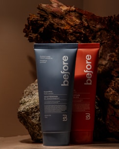 Two toothpaste tubes are positioned against a backdrop of textured, bark-like material. The tube in the foreground is blue and prominently displays the brand name 'before' and the text 'Whitening.' The tube behind it is red with similar branding.