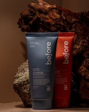 Two toothpaste tubes are positioned against a backdrop of textured, bark-like material. The tube in the foreground is blue and prominently displays the brand name 'before' and the text 'Whitening.' The tube behind it is red with similar branding.