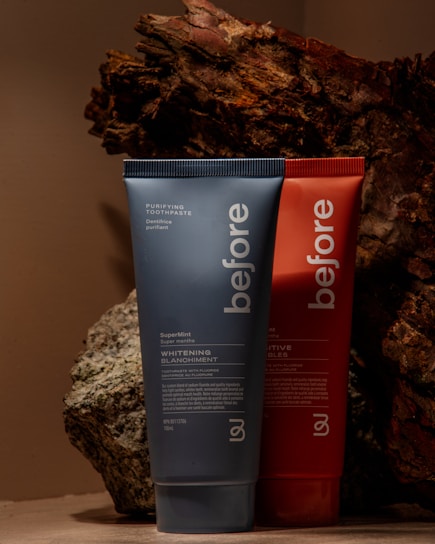 Two toothpaste tubes are positioned against a backdrop of textured, bark-like material. The tube in the foreground is blue and prominently displays the brand name 'before' and the text 'Whitening.' The tube behind it is red with similar branding.