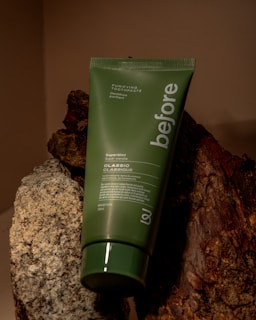 A green tube of 'before' purifying toothpaste labeled SuperMint, placed against a background of rocks and wood. The tube is vertically oriented, partially leaning on the textured surfaces.