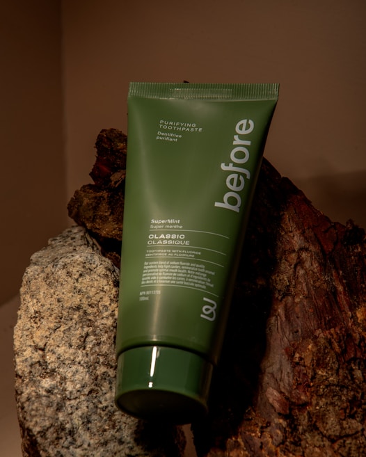 A green tube of 'before' purifying toothpaste labeled SuperMint, placed against a background of rocks and wood. The tube is vertically oriented, partially leaning on the textured surfaces.