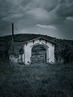 An old, partially dilapidated white archway stands in an overgrown field, with the words 'Fazenda Cruzeiro' inscribed on it. A rustic wooden gate is positioned beneath the arch. The background features rolling hills under a moody, overcast sky.