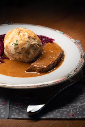 A plate features a serving of beef with rich gravy, accompanied by a large dumpling and a portion of red cabbage. The dish is set on a white plate with an intricate pattern along the edge, placed on a table with a decorative napkin. A fork and spoon are partially visible, adding to the dining ambiance.