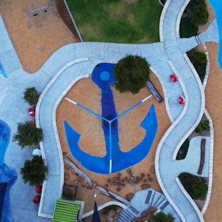 An aerial view of a playground with a large blue anchor design at the center, surrounded by winding pathways and patches of greenery. The layout includes various geometric shapes and textures, such as circular stepping stones and red spherical elements. The area is bordered by trees and grassy sections, creating a harmonious blend of natural and artificial elements.