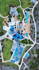 An aerial view of a landscaped recreational area featuring a playground with various play structures, a basketball court with blue markings, walking paths, grassy fields, and scattered trees. Several modern structures with blue roofs are also visible, surrounded by green and brown landscaped areas.