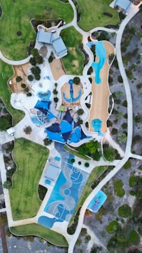 An aerial view of a landscaped recreational area featuring a playground with various play structures, a basketball court with blue markings, walking paths, grassy fields, and scattered trees. Several modern structures with blue roofs are also visible, surrounded by green and brown landscaped areas.