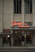 A street view of a restaurant with a prominent neon sign labeled 'Fran's Restaurant' above the entrance. The building is a muted beige with tall windows, and there are glass doors leading into the eatery. To the side, a small outdoor seating area is visible, partially sheltered by an awning, and a warm glow emanates from the interior.