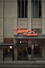 A street view of a restaurant with a prominent neon sign labeled 'Fran's Restaurant' above the entrance. The building is a muted beige with tall windows, and there are glass doors leading into the eatery. To the side, a small outdoor seating area is visible, partially sheltered by an awning, and a warm glow emanates from the interior.