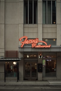 A street view of a restaurant with a prominent neon sign labeled 'Fran's Restaurant' above the entrance. The building is a muted beige with tall windows, and there are glass doors leading into the eatery. To the side, a small outdoor seating area is visible, partially sheltered by an awning, and a warm glow emanates from the interior.