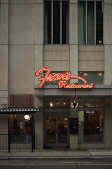 A street view of a restaurant with a prominent neon sign labeled 'Fran's Restaurant' above the entrance. The building is a muted beige with tall windows, and there are glass doors leading into the eatery. To the side, a small outdoor seating area is visible, partially sheltered by an awning, and a warm glow emanates from the interior.