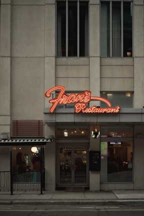 A street view of a restaurant with a prominent neon sign labeled 'Fran's Restaurant' above the entrance. The building is a muted beige with tall windows, and there are glass doors leading into the eatery. To the side, a small outdoor seating area is visible, partially sheltered by an awning, and a warm glow emanates from the interior.