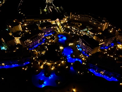 Aerial view of a resort complex at night, featuring illuminated buildings and vividly lit swimming pools with bright blue lights. The scene is filled with warm yellow lighting from various outdoor lights, creating a vibrant and lively atmosphere.