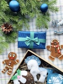 A festive arrangement featuring two gingerbread cookies, a blue wrapped gift box with a green ribbon, a pine cone, and evergreen branches adorned with blue ornaments. Beside the box, there is a decorative silver rocking horse and a selection of felt ornaments in a box, including a white and blue horse and a pudding.