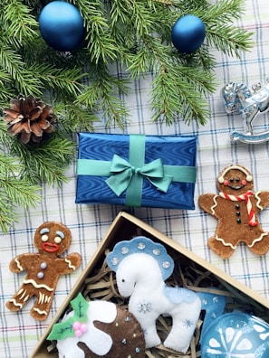 A festive arrangement featuring two gingerbread cookies, a blue wrapped gift box with a green ribbon, a pine cone, and evergreen branches adorned with blue ornaments. Beside the box, there is a decorative silver rocking horse and a selection of felt ornaments in a box, including a white and blue horse and a pudding.
