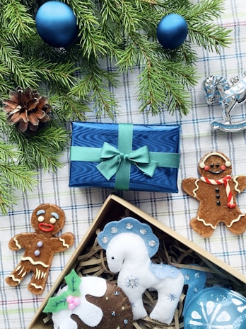 A festive arrangement featuring two gingerbread cookies, a blue wrapped gift box with a green ribbon, a pine cone, and evergreen branches adorned with blue ornaments. Beside the box, there is a decorative silver rocking horse and a selection of felt ornaments in a box, including a white and blue horse and a pudding.