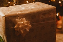 A wrapped Christmas gift featuring light brown paper with cartoon animal designs and a large decorative bow. A tag on the gift reads 'To: grandma, From: Felicia.' The background is softly lit with warm, glowing lights, suggesting a cozy holiday setting.