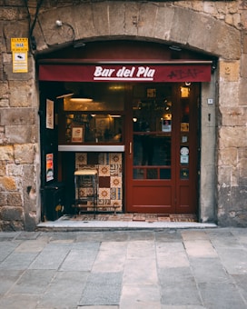 A quaint bar entrance with a rustic stone facade and a maroon canopy displaying the name 'Bar del Pla'. The doorway is framed by a mix of patterned tiles, and a couple of high chairs are placed at the entrance. The warm lighting from inside gives the scene a welcoming ambiance.