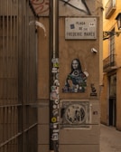 A narrow street with urban art and graffiti. A sign reading 'Plaça de Frederic Marès' is mounted on a wall. Underneath, a modern take on the Mona Lisa covered with colorful tags and stickers is present. A pole in the foreground is adorned with various stickers, adding to the street art vibe. The setting is framed by warm-toned walls and a barred fence to the left, giving an enclosed atmosphere.