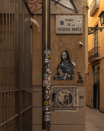 A narrow street with urban art and graffiti. A sign reading 'Plaça de Frederic Marès' is mounted on a wall. Underneath, a modern take on the Mona Lisa covered with colorful tags and stickers is present. A pole in the foreground is adorned with various stickers, adding to the street art vibe. The setting is framed by warm-toned walls and a barred fence to the left, giving an enclosed atmosphere.