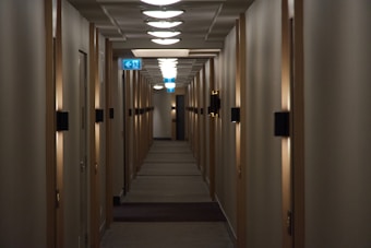 A dimly lit hotel corridor with closed wooden doors on either side. The ceiling is lined with circular lights creating a repeating pattern down the hall. An illuminated exit sign is visible towards the end, indicating the direction of an escape route.