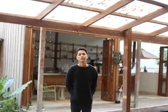 A person stands under a wooden pergola with an open door leading to an indoor space in the background. The setting appears to be a combination of indoor and outdoor areas, featuring wooden structures, a few plants, and a partially visible kitchen or dining area inside.