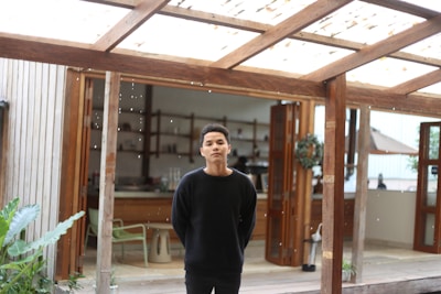A person stands under a wooden pergola with an open door leading to an indoor space in the background. The setting appears to be a combination of indoor and outdoor areas, featuring wooden structures, a few plants, and a partially visible kitchen or dining area inside.