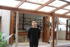 A person stands under a wooden pergola with an open door leading to an indoor space in the background. The setting appears to be a combination of indoor and outdoor areas, featuring wooden structures, a few plants, and a partially visible kitchen or dining area inside.