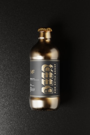 A gold-colored beer bottle with a metallic finish resting against a dark, textured background. The large, stylized word 'BEER' is prominent on the label, along with smaller text and a circular logo above it. The bottle has a short neck with a matching gold cap.