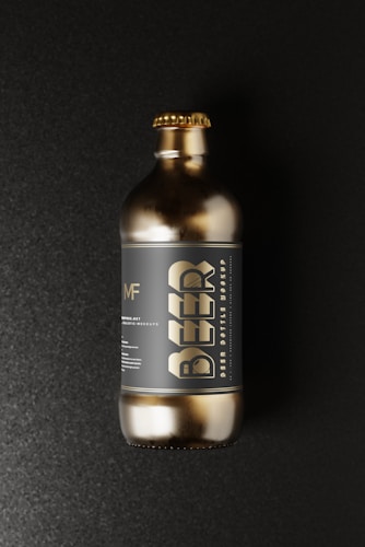 A gold-colored beer bottle with a metallic finish resting against a dark, textured background. The large, stylized word 'BEER' is prominent on the label, along with smaller text and a circular logo above it. The bottle has a short neck with a matching gold cap.