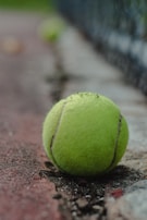 A tennis ball lies on a clay tennis court, with a slightly out-of-focus background. The ball is worn and has dirt marks, and the texture of the court surface is visible.