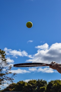 A tennis ball is in mid-air against a backdrop of a clear blue sky, with a few scattered clouds. In the foreground, a hand holds a tennis racket poised to strike the ball. There is also some foliage seen at the lower edges of the image.