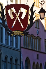 A decorative sign with a large red shield and crossed tools hangs in front of old buildings with arches and a street lamp. The sign has German text that reads 'Die Altstadt' and adds to the historical charm of the scene.