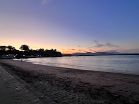A serene beach setting during sunset, with soft colors of dusk in the sky blending from warm yellows to deep blues. Silhouettes of trees and distant hills outline the horizon, while gentle waves lap against the sandy shore. A couple can be seen walking along the beach, adding a peaceful human element to the tranquil scene.