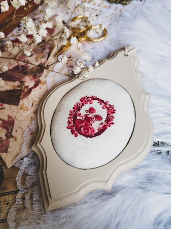 A vintage-style embroidery hoop framed in ornate beige wood is placed on a soft, textured fabric. The embroidery features a detailed red floral pattern on white fabric. Surrounding the frame, delicate dried flowers and golden scissors add a touch of elegance, while sheets of lace and musical notes create a nostalgic atmosphere.