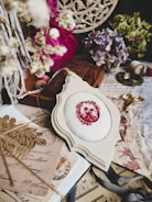A vintage-inspired arrangement of crafting and decorative items. The centerpiece is a piece of fabric with an embroidered red floral pattern, surrounded by dried flowers, an antique lace cloth, a pair of golden scissors, ribbon, spools of thread, old postcards, and sheet music. The color palette includes soft pastels and faded hues, creating a nostalgic atmosphere.