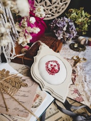 A vintage-inspired arrangement of crafting and decorative items. The centerpiece is a piece of fabric with an embroidered red floral pattern, surrounded by dried flowers, an antique lace cloth, a pair of golden scissors, ribbon, spools of thread, old postcards, and sheet music. The color palette includes soft pastels and faded hues, creating a nostalgic atmosphere.