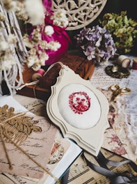 A vintage-inspired arrangement of crafting and decorative items. The centerpiece is a piece of fabric with an embroidered red floral pattern, surrounded by dried flowers, an antique lace cloth, a pair of golden scissors, ribbon, spools of thread, old postcards, and sheet music. The color palette includes soft pastels and faded hues, creating a nostalgic atmosphere.