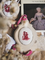 A vintage-style arrangement featuring embroidered fabric with red figures in an ornate frame. A historical portrait of a woman in an elegant dress is partially visible beneath the frame. Dried flowers are blurred in the foreground, adding a rustic touch.
