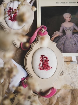 A vintage-style arrangement featuring embroidered fabric with red figures in an ornate frame. A historical portrait of a woman in an elegant dress is partially visible beneath the frame. Dried flowers are blurred in the foreground, adding a rustic touch.
