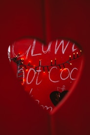 Through a heart-shaped cutout, a red backdrop with words in white chalk is visible, partially obscured by a string of small, glowing yellow lights, creating a warm and intimate setting.