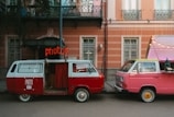 Two vintage vans are parked side by side, one red and one pink. The red van has an open door with a red curtain inside, functioning as a photo booth with a bright neon sign reading 'photos' above it. The pink van has a pop-up roof adorned with string lights. They are parked in front of a peach-colored building with white-framed windows and balconies.
