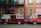 Two vintage vans are parked side by side, one red and one pink. The red van has an open door with a red curtain inside, functioning as a photo booth with a bright neon sign reading 'photos' above it. The pink van has a pop-up roof adorned with string lights. They are parked in front of a peach-colored building with white-framed windows and balconies.
