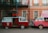 Two vintage vans are parked side by side, one red and one pink. The red van has an open door with a red curtain inside, functioning as a photo booth with a bright neon sign reading 'photos' above it. The pink van has a pop-up roof adorned with string lights. They are parked in front of a peach-colored building with white-framed windows and balconies.