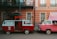 Two vintage vans are parked side by side, one red and one pink. The red van has an open door with a red curtain inside, functioning as a photo booth with a bright neon sign reading 'photos' above it. The pink van has a pop-up roof adorned with string lights. They are parked in front of a peach-colored building with white-framed windows and balconies.