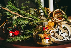 Holiday-themed decorations with clear glass baubles containing small mushrooms and glittery stars, surrounded by green pine branches. A wooden heart ornament with a cut-out reindeer is also visible.