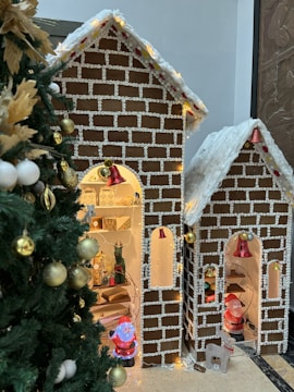 A Christmas-themed setup featuring two gingerbread house decorations adorned with white icing patterns and yellow lights. The houses are detailed with small red bells and contain Christmas figures inside. To the left, a decorated Christmas tree is partially visible with gold and white ornaments.