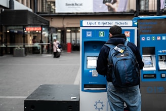 A person with a backpack is using a blue ticket vending machine in a public outdoor area with shops and signage in the background.