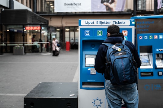 A person with a backpack is using a blue ticket vending machine in a public outdoor area with shops and signage in the background.