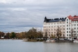 A waterfront scene featuring a row of elegant multi-story buildings with intricate architectural designs. The buildings have varied roof colors including black and red, and sit along a calm body of water. Leafless trees stand in front of the buildings, indicating an autumn setting. Small boats are docked near the shoreline.